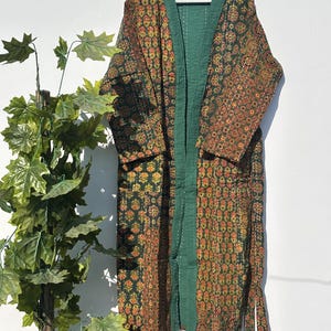 Pode incluir: Robe comprido, aberto na frente, com interior e acabamentos verdes, e exterior estampado em tons de verde, laranja e amarelo. O robe está pendurado num cabide de madeira contra uma parede branca. O tecido parece acolchoado.