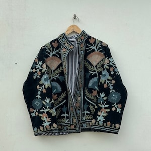 Puede incluir: Chaqueta negra bordada con diseños florales y de hojas en tonos azules, melocotón y verdes. La chaqueta tiene cuello mandarín y forro a rayas. La chaqueta está colgada en una percha de madera.