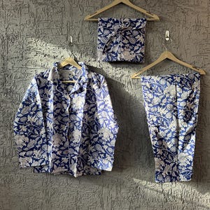 Può includere: Un completo pigiama floreale blu e bianco, composto da una camicia con bottoni, pantaloni e un top piegato, tutti appesi a grucce di legno. Il tessuto presenta un motivo ripetuto di fiori e foglie bianche su sfondo blu navy.