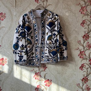 Può includere: Giacca bianca con ricami floreali intricati in blu, oro e nero. La giacca presenta un colletto alla coreana ed è appesa a un'appendiabiti di legno. Il design include motivi floreali dettagliati, creando un aspetto decorativo e artistico.