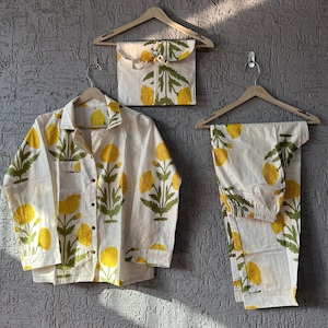 Op de afbeelding: Witte pyjamaset met een geel bloemmotief, bestaande uit een overhemd met knopen, een broek en een bijpassend etui. Het bloemmotief toont gele bloemen en groene bladeren. De set wordt gepresenteerd op houten kleerhangers.