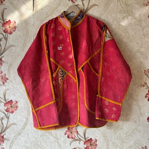 Peut inclure: Veste matelassée rouge avec garniture jaune et détails patchwork. La veste est suspendue à un cintre en bois sur un fond de papier peint floral. La veste a un col et une fermeture à boutons.