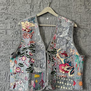 Puede incluir: Un chaleco acolchado con un estampado floral y de dragón en tonos grises, rosas y verdes. El chaleco tiene un forro y ribete a rayas blancas y negras. Es sin mangas y cuelga de una percha de madera.