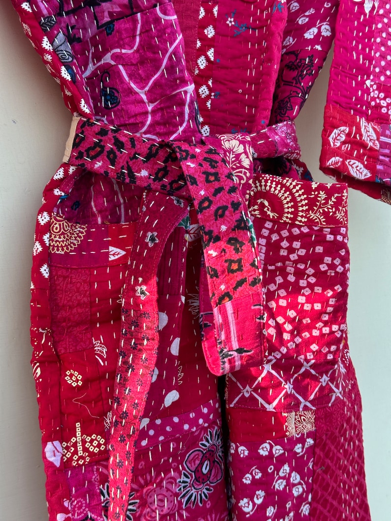 Pode incluir: Robe de patchwork em tons de vermelho e rosa, com v&aacute;rios padr&otilde;es florais e geom&eacute;tricos. O robe tem um cinto a condizer com um design de padr&atilde;o de leopardo. As costuras brancas detalham as costuras e os padr&otilde;es, criando um visual texturizado.