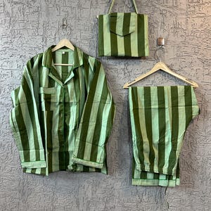 May include: A green striped pajama set and matching tote bag. The set includes a button-down shirt and pants, all hanging on wooden hangers. The tote bag has a flap closure and a long strap. The background is a textured gray wall.