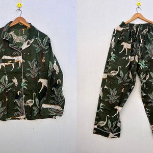May include: Dark green pajama set featuring a jungle animal print. The long-sleeved button-down shirt and pants display zebras, leopards, and birds among stylized trees. White trim outlines the collar, cuffs, and pockets. The set is hanging on a wooden hanger.