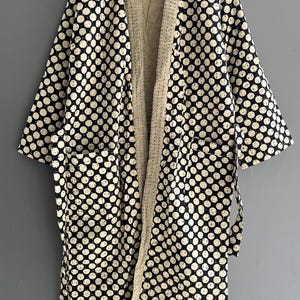 May include: A black and white patterned kimono-style robe with a cream-colored interior. The robe features a repeating polka dot design and two front pockets. The robe is hanging on a wooden hanger against a gray background.