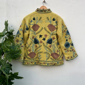 May include: A yellow jacket with detailed floral embroidery in brown, blue, and green. The jacket has a textured, waffle-like pattern and a high collar. The back of the jacket features a large floral design.