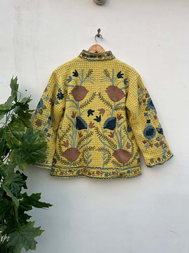 May include: A yellow jacket with detailed floral embroidery in brown, blue, and green. The jacket has a textured, waffle-like pattern and a high collar. The back of the jacket features a large floral design.