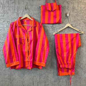 Puede incluir: Un conjunto de pijama y un bolso tote a juego con un vibrante estampado de rayas rosas y naranjas. El conjunto incluye una camisa abotonada, pantalones y un bolso tote con cierre de solapa. El conjunto de pijama está colgado en una percha de madera contra una pared gris texturizada.