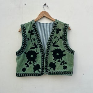 以下が含まれることがあります： 黒い花の刺繍とトリムが施されたセージグリーンのベスト。ノースリーブのベストにはストライプの裏地が付いています。刺繍は、大きく様式化された花と葉の模様が特徴です。木製のハンガーにかかっています。