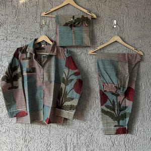 Peut inclure: Un ensemble de pyjama assorti avec un imprimé floral rouge, turquoise et vert. L'ensemble comprend une chemise boutonnée, un pantalon et une taie d'oreiller assortie, le tout suspendu à des cintres en bois sur un mur gris texturé.