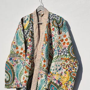 May include: A colorful, quilted robe with a paisley and floral pattern. The robe features a cream-colored base with vibrant accents of blue, orange, green, and yellow. The robe is hanging on a metal hanger.