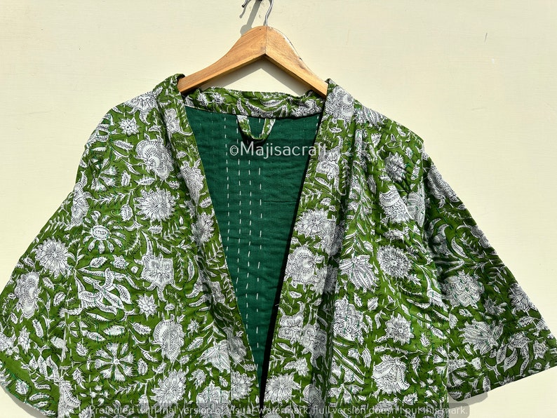 Może przedstawiać: Zielono-biała kurtka w stylu kimono z kwiatowym nadrukiem. Kurtka z otwartym przodem ma ciemnozielone wnętrze z białymi przeszyciami. Wz&oacute;r kwiatowy to powtarzający się motyw liści i kwiat&oacute;w.