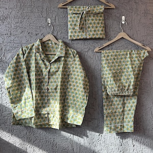 Può includere: Un completo pigiama verde chiaro con motivo floreale, composto da una camicia con bottoni, pantaloni e una borsa con coulisse abbinata, il tutto appeso a grucce di legno contro un muro grigio strutturato.