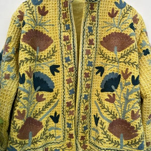 May include: A yellow embroidered jacket with a waffle-weave texture. The jacket features a floral design in shades of brown, blue, and green. The collar and edges are also embroidered with a similar floral pattern. The jacket is open in the front.