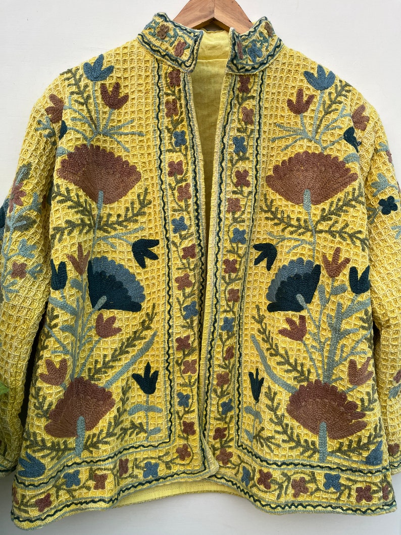May include: A yellow embroidered jacket with a waffle-weave texture. The jacket features a floral design in shades of brown, blue, and green. The collar and edges are also embroidered with a similar floral pattern. The jacket is open in the front.