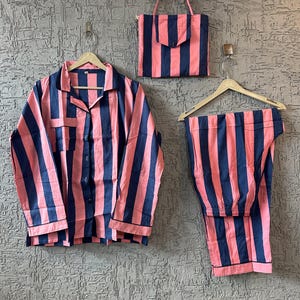Puede incluir: Un conjunto de pijama y bolso a juego con un estampado de rayas rosas y azul marino. El conjunto incluye una camisa abotonada, pantalones y un bolso con un bolsillo. Los pijamas están colgados en perchas de madera.