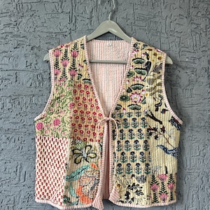 Puede incluir: Un chaleco acolchado con un diseño de patchwork con estampados florales y botánicos en tonos rosa, rojo, verde y azul sobre un fondo crema. El chaleco tiene un cierre de lazo y un forro a rayas rosas.