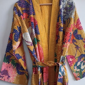 Puede incluir: Bata tipo kimono de color amarillo mostaza con un estampado floral en rosa, rojo, morado y azul. La bata tiene un cinturón a juego y costuras blancas. Confeccionada con un tejido suave y ligero.