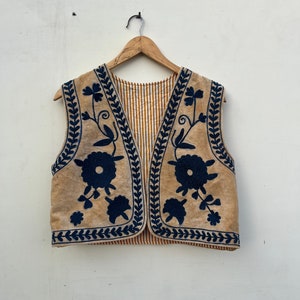 May include: A tan vest with dark blue floral embroidery and trim. The vest has a striped brown and white lining and is hanging on a wooden hanger against a white wall. The embroidery features stylized flowers and vines.