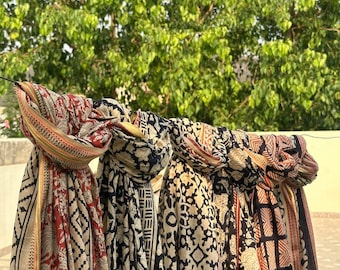 May include: Several patterned scarves hang outdoors. The scarves feature various designs in red, black, and beige. The patterns include geometric shapes, floral motifs, and traditional block prints. The scarves are draped over a line against a backdrop of green foliage.