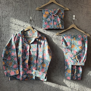 May include: A blue floral pajama set, comprising a button-down shirt and trousers, hangs on a wooden hanger. The set features a pink and yellow floral pattern. A matching set, folded and tied with a ribbon, is also visible.