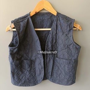 May include: A navy blue vest with two front pockets and a V-neck design. The vest is made of a textured fabric and hangs on a wooden hanger. The brand name "Majisacraft" is visible on the vest.