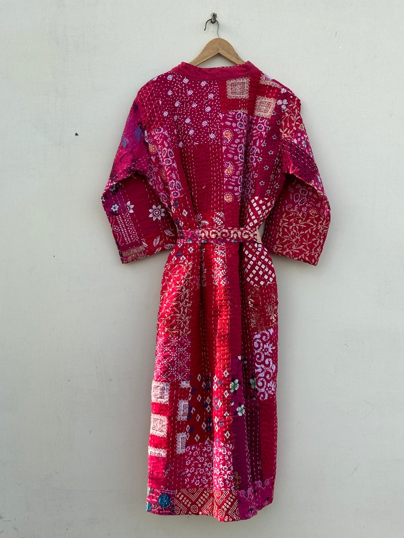 Pode incluir: Um roup&atilde;o acolchoado de patchwork em tons de vermelho e rosa est&aacute; pendurado em um cabide de madeira. O roup&atilde;o apresenta uma variedade de quadrados de tecido estampado, com um cinto a condizer. &Eacute; uma pe&ccedil;a longa e fluida.