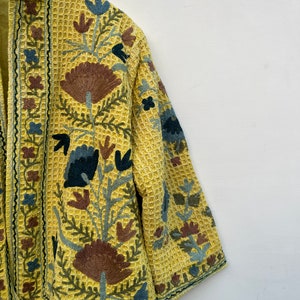 May include: A yellow jacket with detailed floral embroidery in brown, blue, and green. The jacket has a textured, waffle-like fabric and a mandarin collar. The embroidery is intricate, covering the front and sleeves.
