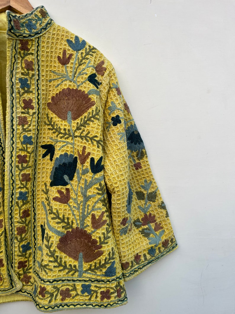 May include: A yellow jacket with detailed floral embroidery in brown, blue, and green. The jacket has a textured, waffle-like fabric and a mandarin collar. The embroidery is intricate, covering the front and sleeves.