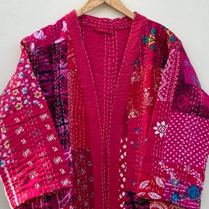 Pode incluir: Um quimono patchwork em tons de rosa e vermelho, com padr&otilde;es florais e geom&eacute;tricos. O roup&atilde;o &eacute; feito de tecido costurado e pendurado em um cabide de madeira. A pe&ccedil;a tem decote em V e mangas compridas.