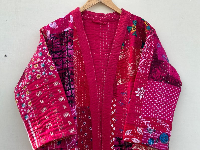 Pode incluir: Um quimono patchwork em tons de rosa e vermelho, com padr&otilde;es florais e geom&eacute;tricos. O roup&atilde;o &eacute; feito de tecido costurado e pendurado em um cabide de madeira. A pe&ccedil;a tem decote em V e mangas compridas.