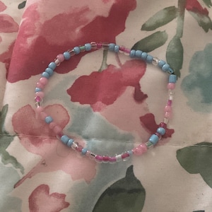 Op de afbeelding: Een delicate armband gemaakt met kleine roze en blauwe kralen. De armband ligt op een stoffen achtergrond met een bloemenpatroon.