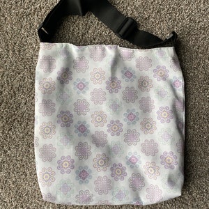 May include: A white fabric tote bag with a black adjustable shoulder strap. The bag has a floral pattern with purple, blue, and yellow swirls.