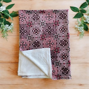 May include: A soft, plush blanket with a pink and brown geometric pattern. The blanket is folded in half, showing the white fleece lining.