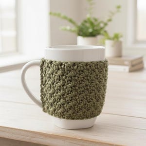May include: A white ceramic mug with a green crocheted cozy. The cozy is a solid olive green color and covers the middle section of the mug. The mug is on a light-colored wooden surface.