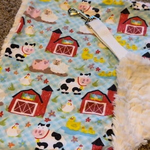 May include: A blue and white fabric baby blanket with a farm animal theme. The blanket features cows, pigs, chickens, ducks, and red barns. The blanket has a white, fluffy trim around the edges.