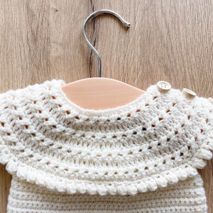 May include: A white crocheted baby garment with a scalloped collar and two wooden buttons. The garment is hanging on a wooden hanger.