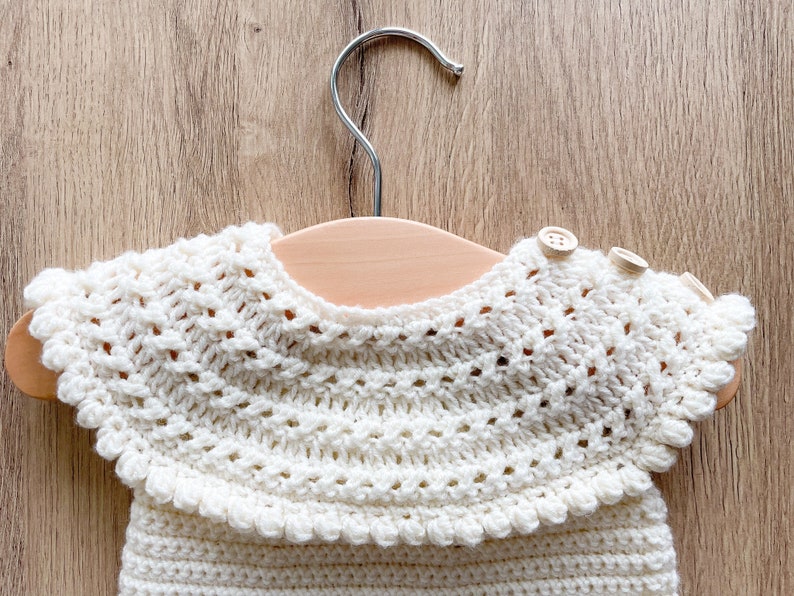 May include: A white crocheted baby garment with a scalloped collar and two wooden buttons. The garment is hanging on a wooden hanger.