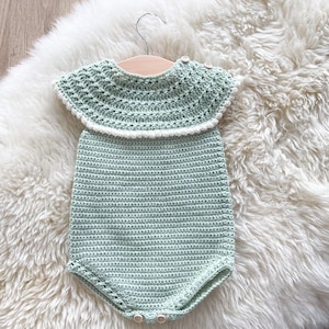 May include: A crocheted baby onesie in a light green color with a white crocheted collar. The onesie is hanging on a wooden hanger on a white fluffy surface.