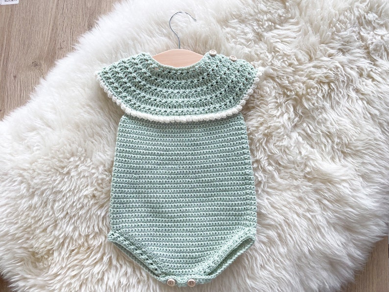 May include: A crocheted baby onesie in a light green color with a white crocheted collar. The onesie is hanging on a wooden hanger on a white fluffy surface.