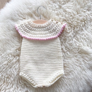 May include: A white crocheted baby onesie with a pink crocheted trim around the neckline. The onesie has a button closure at the bottom.
