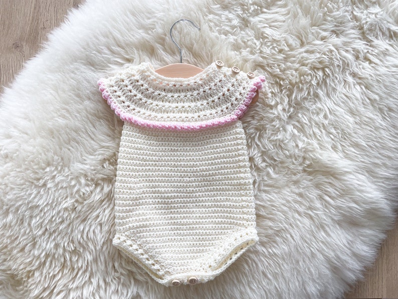 May include: A white crocheted baby onesie with a pink crocheted trim around the neckline. The onesie has a button closure at the bottom.