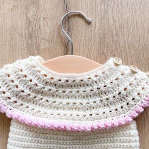May include: A white crocheted baby garment with a pink trim and wooden buttons. The garment is hanging on a wooden hanger.