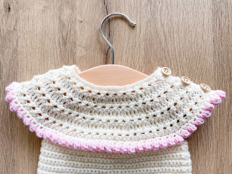 May include: A white crocheted baby garment with a pink trim and wooden buttons. The garment is hanging on a wooden hanger.