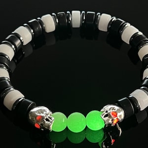 Pode incluir: Pulseira de contas pretas e brancas com berloques de caveira prateados com detalhes vermelhos e três contas verdes brilhantes. As contas cilíndricas alternam preto e branco.