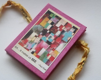Puede incluir: Un pequeño objeto rectangular rosa con una colorida impresión de arte abstracto. La impresión presenta un mosaico de cuadrados en rojo, azul, amarillo y verde. El texto en el objeto dice "Art... It makes us SEE." y "-Paul Klee."