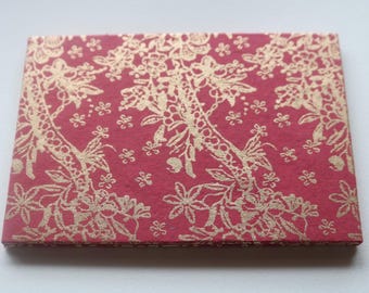 Puede incluir: Caja rectangular cerrada con fondo rojo y un patrón de encaje floral dorado. La caja, probablemente de papel o cartulina, es adecuada para pequeños regalos o joyas.