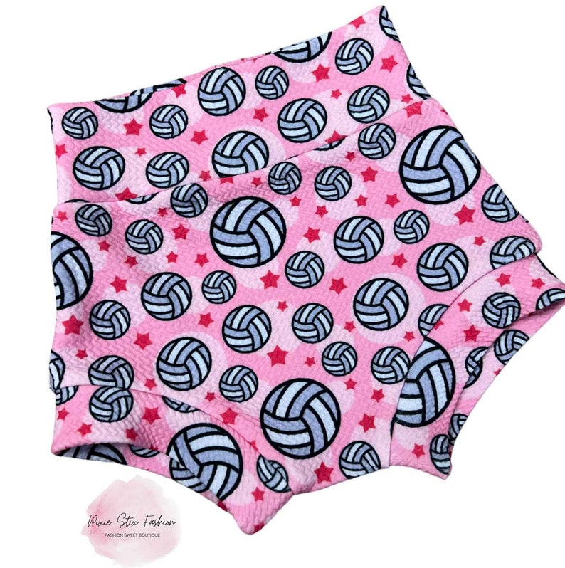 May include: Pink high-waisted shorts with a repeating pattern of volleyballs and red stars. The volleyballs are white and light blue with black outlines. The shorts have a textured fabric and a high waistband.